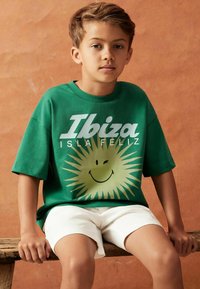 Camiseta verde de algodón con el texto "Ibiza" y un gráfico de un sol sonriente, combinada con pantalones cortos blancos. Modelo sentado en un banco de madera.