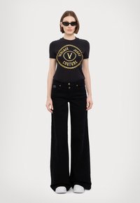 Black cotton t-shirt with gold logo, paired with high-waisted black wide-leg jeans, and white slip-on shoes. Accessories include black sunglasses.