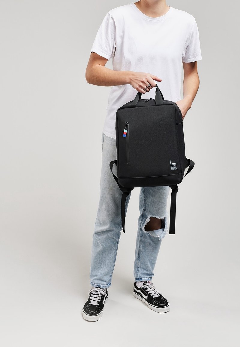 Sac à dos noir en tissu résistant, avec une poche avant zippée, des bretelles réglables et une petite étiquette tricolore en accent.