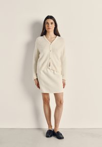 Krémově zbarvený, huňatý pletený kardigan s výstřihem do V a knoflíkovým zapínáním, spárovaný s bílou sukní a černými mokasíny.