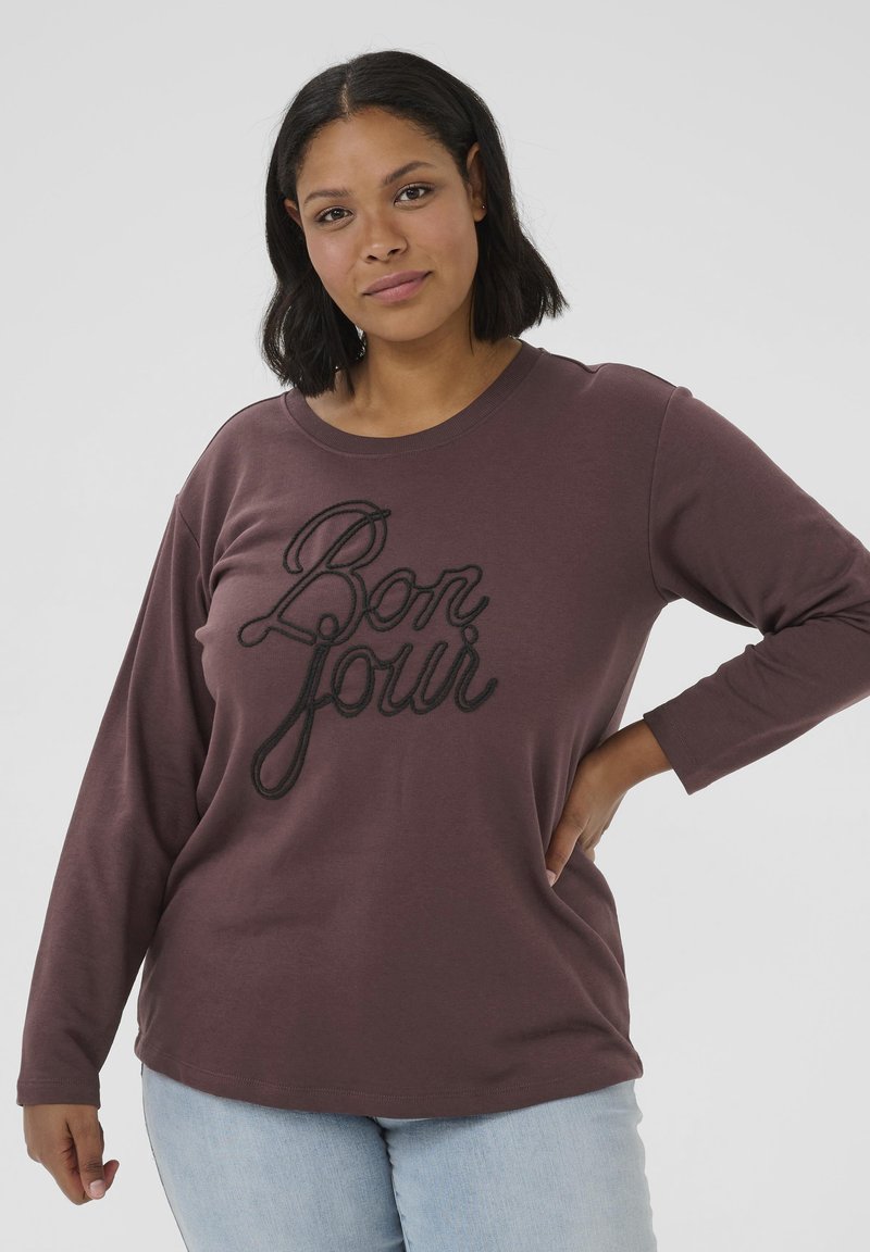 Chemise marron à manches longues en tissu doux avec "Bon Jour" brodé en cursive noire. La chemise a une coupe décontractée et un col rond.