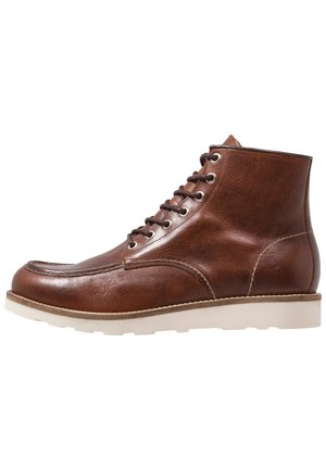 Bottines en cuir marron avec une texture lisse, bout rond, six œillets en métal et une semelle en caoutchouc blanc. Détails cousus sur les coutures.