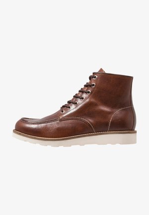 Bottines en cuir marron avec une texture lisse, bout rond, six œillets en métal et une semelle en caoutchouc blanc. Détails cousus sur les coutures.
