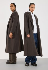 Brown double-breasted long coat with large lapels, displayed on two models. One wears a white hoodie and the other blue jeans. Textured fabric.
