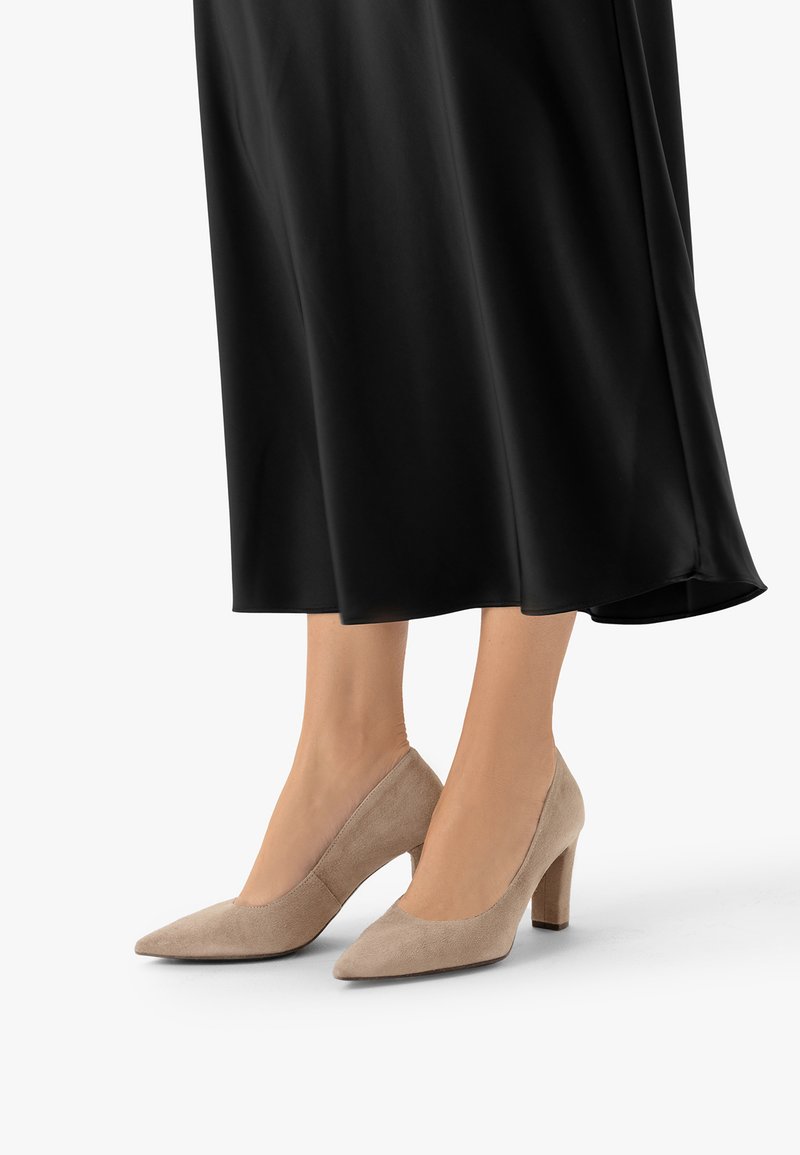 Jambes portant des chaussures beige en daim à bout pointu et talon moyen, avec une jupe noire fluide sur un fond clair.