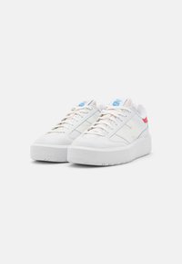 Sneakers bianchi con una texture liscia, dotati di un collare imbottito, accenti blu e rossi, e una suola in gomma spessa. Posizionati uno accanto all'altro.