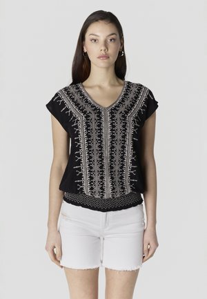 Mujer joven con cabello largo y oscuro, vestida con blusa negra bordada y pantalones cortos de mezclilla blancos, de pie contra un fondo liso.