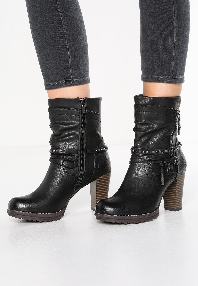 Bottes en cuir noir mi-mollet avec fermetures éclair latérales, sangles décoratives et talons blocs empilés portées avec un pantalon ajusté foncé.
