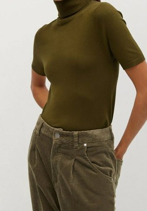 Kvinde iført en olivengrøn kortærmet rullekravstopp og brune, højtaljede bukser af fløjl med hænderne i lommerne.