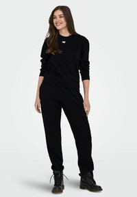 Νεαρή γυναίκα με μακριά καστανά μαλλιά, ντυμένη με μαύρο φούτερ, μαύρο παντελόνι, μαύρες μποτίνες με κορδόνια και ένα κολιέ, χαμογελώντας και στέκεται.