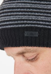 Sort og grå stribet strikbeanie med ribbet tekstur. Har et sort logomærke på kanten. Nærbillede af hatten på et hoved.