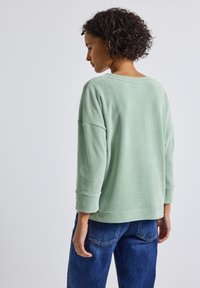 Lichtgroene, gestructureerde, lange mouwen trui met een relaxed fit en opgerolde manchetten, gecombineerd met blauwe jeans, van achteren weergegeven.