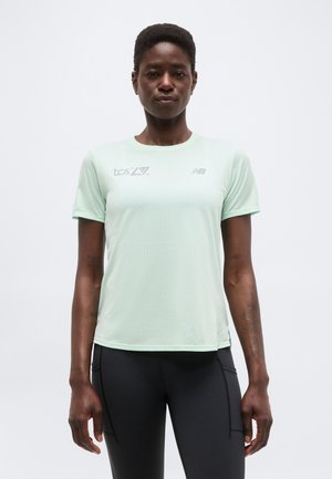 Γυναίκα που φοράει αθλητική μπλούζα κοντό μανίκι ανοιχτού πράσινου χρώματος με λογότυπα και μαύρο κολάν, στέκεται μπροστά σε απλό λευκό φόντο.