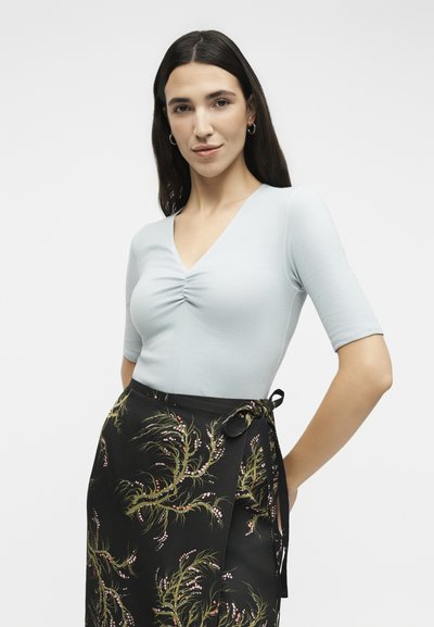 Γυναίκα με μακριά σκούρα μαλλιά, που φοράει ανοιχτό μπλε μπλουζάκι με κοντά μανίκια και μαύρη φλοράλ φούστα τύπου wrap, στέκεται με τα χέρια στη μέση σε λευκό φόντο.