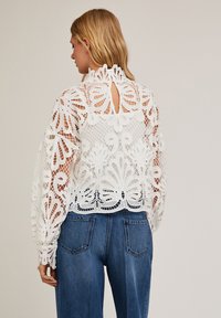 Witte kanten blouse met ingewikkelde bloemmotieven, hoge hals en sleutelgatrug; in combinatie met blauwe spijkerbroeken. Getextureerde en lichte stof.