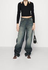 Schwarzes geripptes Croptop mit ausgeschnittenem Kragen, kombiniert mit lockeren blauen Jeans und braunen Sneakers. Eine schwarze Handtasche mit Kettendetails wird gehalten.