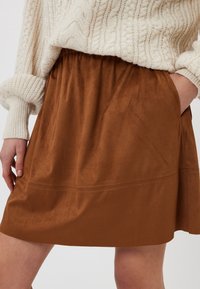 Jupe mini en daim marron avec une taille élastique et des poches latérales, présentant des détails de couture discrets le long de l'ourlet.