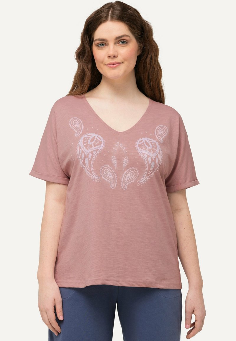 Camiseta de manga corta en rosa claro con escote en V, que presenta un estampado de paisley blanco y un corte holgado. Tela suave con una textura lisa. Combinada con pantalones azules.