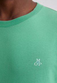 Zielona bawełniana koszulka z okrągłym dekoltem, z białym haftowanym logo „M O'P” na klatce piersiowej. Gładka faktura, dopasowany krój.