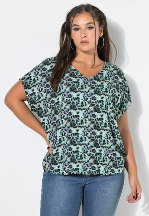 Kvinde med langt hår og øreringe i form af ringe, iført en grøn leopardprint V-hals bluse og blå jeans mod en ensfarvet hvid baggrund.