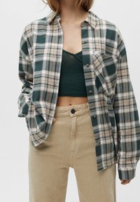 Vrouw in een groene crop top onder een open groen en beige geruit overhemd met beige high-waisted broek.