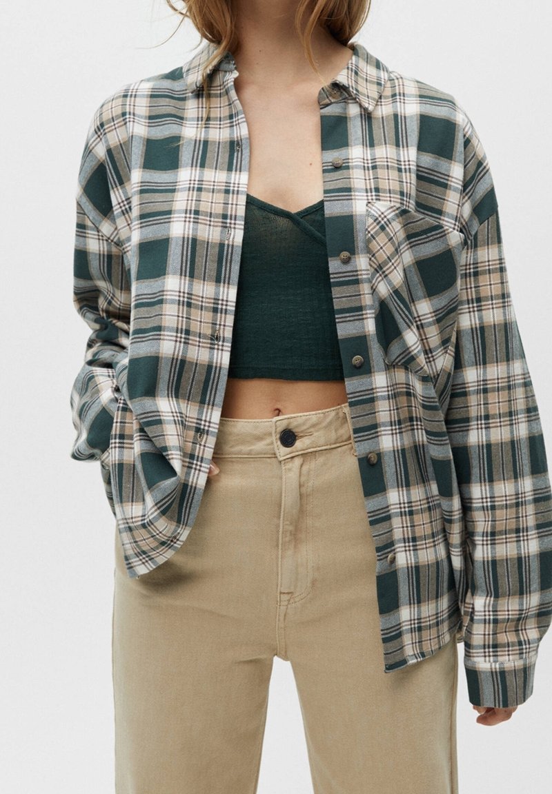 Eine Frau trägt ein grünes Crop-Top unter einem offenen, grün-beigen karierten Hemd und beigen hoch taillierten Hosen.