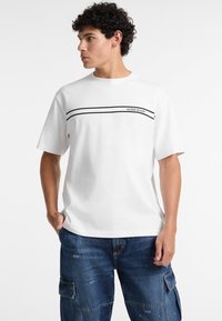 Bjela pamučna majica s kratkim rukavima, s crnim horizontalnim prugama i logotipom "GUESS JEANS" u sredini. Ležeran kroj.