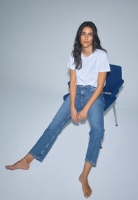 Balta īsroku T-krekls un vidēji zilas taisnas bikses. Modelis sēž uz zilas krēsla, demonstrējot vienkāršu, neformālu izskatu.