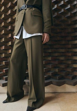 Osoba ubrana w oliwkowo-zielone szerokie spodnie garniturowe i dopasowaną marynarkę z paskiem, na białej koszuli, stojąca na tle teksturowanej ściany.