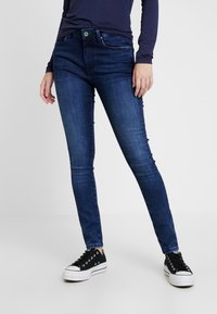 Hubené džíny v tmavě modrém denimu, s klasickým pětihlavým designem, minimálním prošitím a zapínáním na knoflík. Vyznačují se černými teniskami.