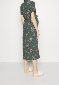 Abito midi floreale verde con maniche corte e vita legata. Presenta fiori rosa brillante e blu. Indossato con zoccoli bianchi con tacco.