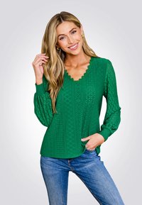 Maglietta verde a maniche lunghe con scollatura a V scallopata, motivo intricata e tessuto testurizzato, abbinata a jeans blu.