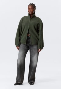 Femeie în picioare purtând o jachetă verde de fleece cu fermoar, top crop negru, blugi largi decolorate și pantofi negri cu vârf ascuțit pe un fundal alb.