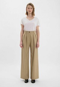 Pantalons beiges à jambes larges avec une taille haute, deux boutons et des plis ; associés à un T-shirt blanc à col en V et des chaussures noires.
