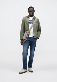 Μπουφάν ελιάς με καφέ γιακά, λευκό πουκάμισο με γραφικό, μπλε τζιν, γκρι τσάντα χιαστί και μπεζ αθλητικά παπούτσια με καφέ σόλες.