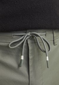 Pantalons vert olive avec un haut noir texturé, présentant un cordon de serrage gris clair avec des embouts verts et blancs fixé devant.