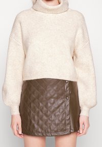 Beżowy sweter z prążkowanym golfem i długimi balonowymi rękawami, zestawiony z brąską, pikowaną skórzaną mini spódnicą z detalami guzików.