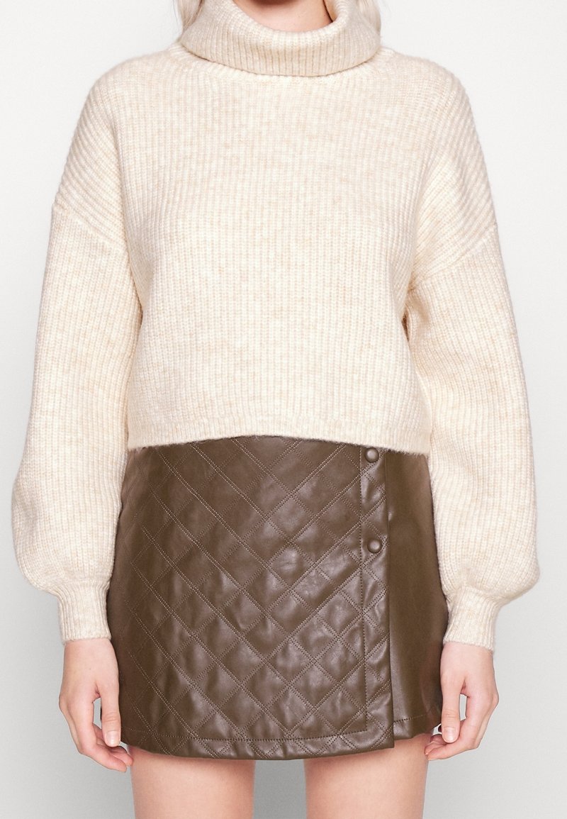 Beżowy sweter z prążkowanym golfem i długimi balonowymi rękawami, zestawiony z brąską, pikowaną skórzaną mini spódnicą z detalami guzików.