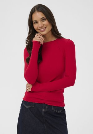 Lächelnde Frau mit langen dunklen Haaren, die ein rotes, geripptes Langarmshirt und einen dunklen Jeansrock trägt, posiert mit der Hand in der Nähe ihres Gesichts.