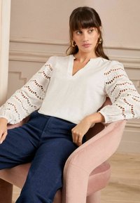 White blouse with eyelet details, deep V-neck, and puffed sleeves, paired with dark blue pants. Seated on a pink upholstered chair.