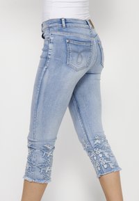 Light blue denim capri jeans with frayed hems and floral embroidery at the cuffs. Features a fitted design and standard back pockets.