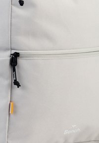 Lichtgrijze rugzak van textuurstof, met een voorste ritsvak, een trekkoord met een togglesluiting en een klein oranje label. Logo gedrukt aan de zijkant.