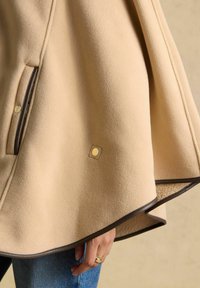 Smėlio spalvos vilnonis pelerinos tipo apsiaustas su tamsiai ruda apvadais ir auksiniais spaustukais, dėvimas ant mėlynų džinsų, matoma ranka su auksiniais žiedais.