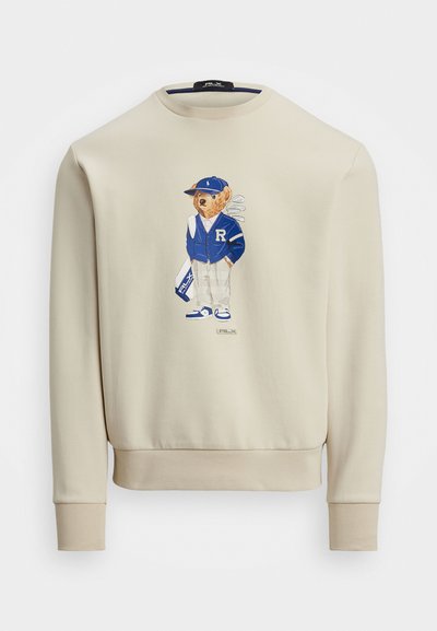 Sweat-shirt beige avec un graphisme d'un ours en veste bleue, tenant des clubs de golf, avec des poignets côtelés et un col rond.