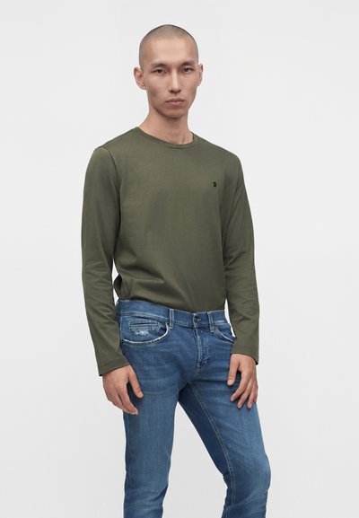 Homme portant une chemise à manches longues vert olive et un jean bleu, debout devant un fond clair uni avec une expression neutre.
