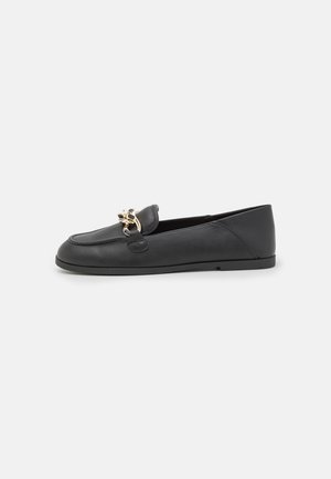 Mocassin plat en cuir noir avec un détail de chaîne dorée sur le dessus, bout arrondi et petit talon sur fond blanc.