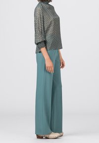 Blusa verde a fantasia con collo alto, abbinata a pantaloni larghi color teal. Scarpe con piattaforma in legno chiaro decorate con borchie. Sfondo neutro.