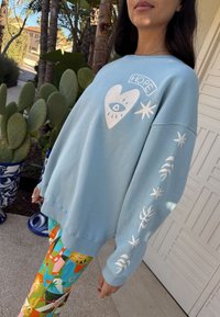 Felpa azzurro chiaro con grafica bianca di cuore e occhio, testo "HOPE" e motivi a foglia bianchi sulle maniche. Abbinata a leggings con motivo colorato.