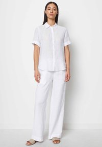 Camicia bianca in lino con maniche corte arrotolate, abbinata a pantaloni larghi in lino bianco e sandali a scivolo color cuoio.