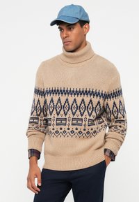 Pull en maille beige à col roulé avec un motif géométrique bleu marine. Manches longues avec un bord côtelé. Porté avec une casquette en denim clair.